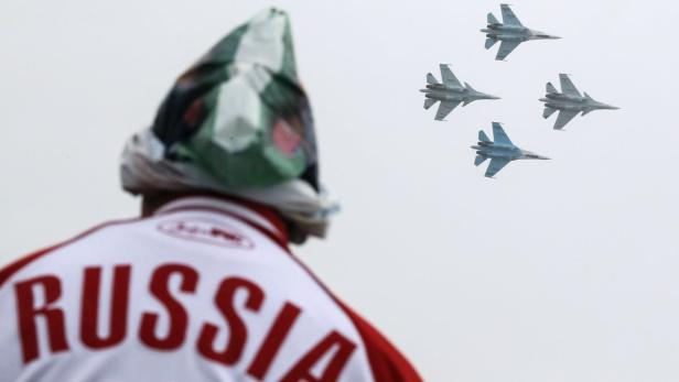 Ein Mann mit Russland-Jacke und Mütze beobachtet vier Kampfflugzeuge am Himmel.