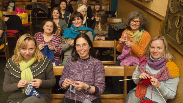 Eine Gruppe Frauen sitzt in einer Reihe und strickt bunte Kleidungsstücke.