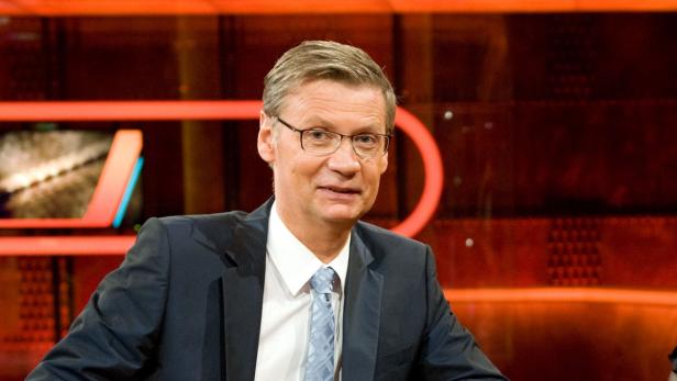 Ein Mann mit Brille und Anzug sitzt vor einem roten Hintergrund.