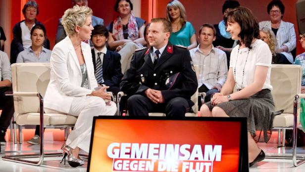 Eine Frau im weißen Hosenanzug im Gespräch mit einem Feuerwehrmann und einer weiteren Frau in einer Fernsehsendung zum Thema Flut.