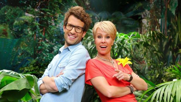 Ein Mann mit Brille und eine Frau mit gelber Blüte stehen Rücken an Rücken vor Dschungelkulisse.