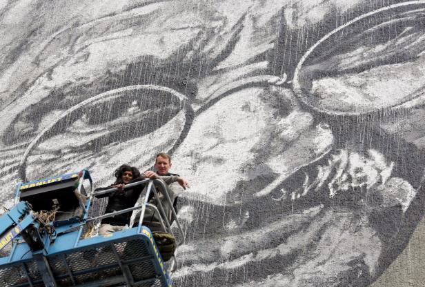 Zwei Personen stehen in einer Hebebühne vor einem großen Wandgemälde.