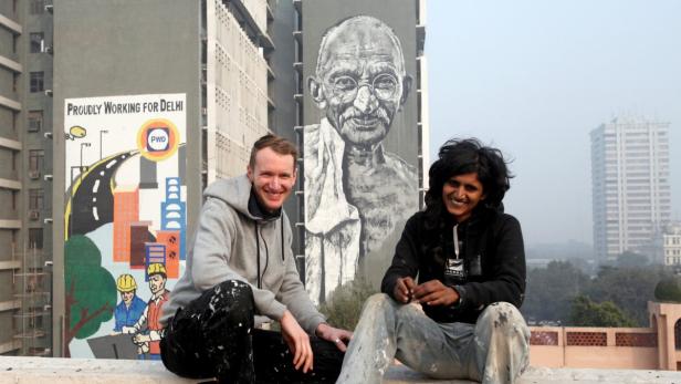 Zwei Künstler sitzen vor einem Gebäude mit einem großen Gandhi-Wandbild in Delhi.