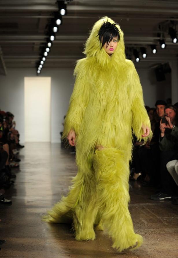 Ein Model präsentiert ein auffälliges, grünes Fellkostüm mit Kapuze auf dem Laufsteg.