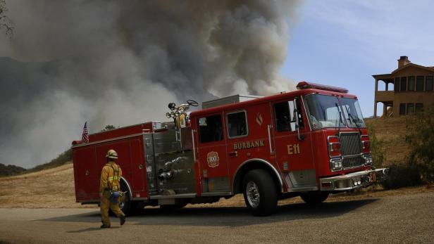 Ein Feuerwehrmann geht an einem roten Feuerwehrwagen der Marke Burbank vorbei, im Hintergrund Rauch.