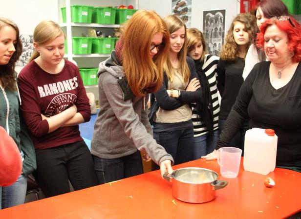 Eine Gruppe von Schülerinnen beobachtet, wie eine rothaarige Schülerin einen Topf auf einen Tisch stellt.