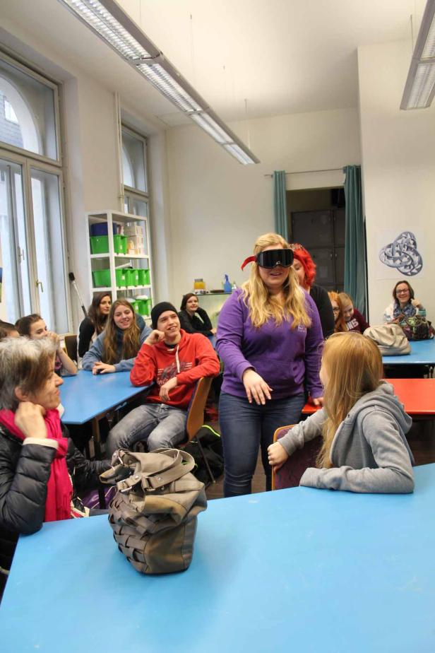 Eine Frau mit verbundenen Augen steht vor einer Gruppe von Menschen in einem Klassenzimmer.