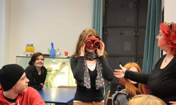 Eine junge Frau setzt eine rote Schutzbrille auf, während andere Personen um einen Tisch sitzen.