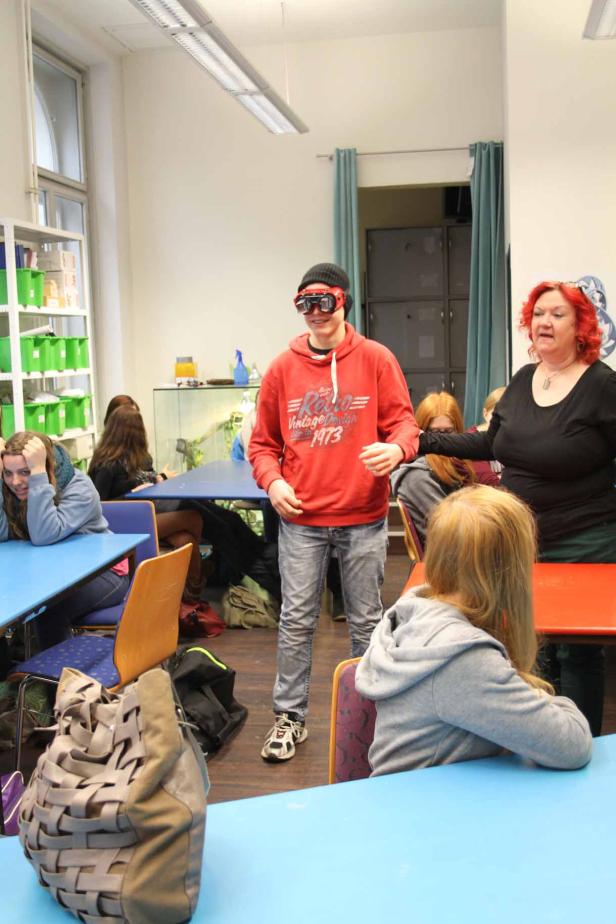 Ein Jugendlicher mit Schutzbrille steht in einem Klassenzimmer, während eine Frau daneben steht.