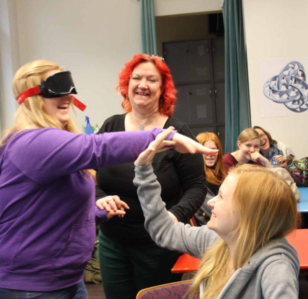 Eine junge Frau mit verbundenen Augen versucht, die Hand einer anderen jungen Frau zu berühren, während andere zusehen.