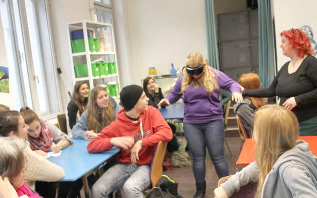 Eine Frau mit verbundenen Augen tastet sich in einem Klassenzimmer voran, während andere Schüler zusehen.