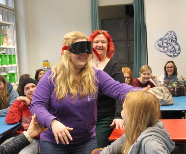 Eine junge Frau mit Skibrille ertastet eine andere junge Frau in einem Klassenzimmer.