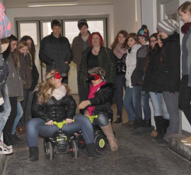 Zwei Frauen mit verbundenen Augen sitzen auf Spielzeugautos, umgeben von einer Gruppe von Zuschauern.