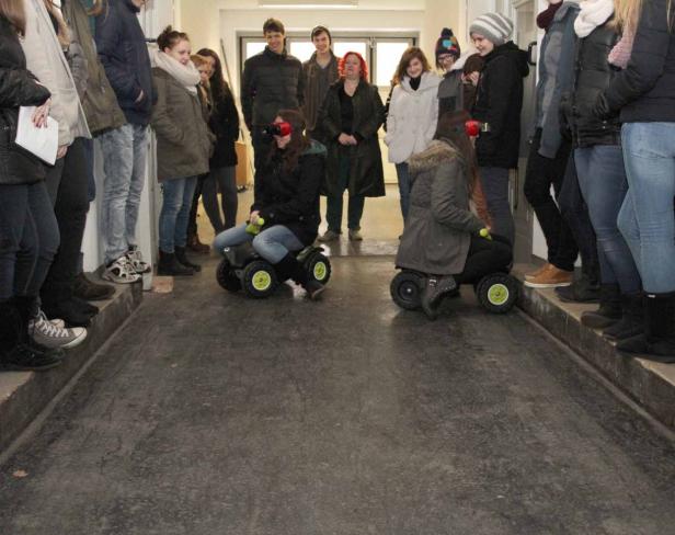 Zwei Frauen fahren auf kleinen Spielzeugautos einen Flur entlang, während andere zusehen.