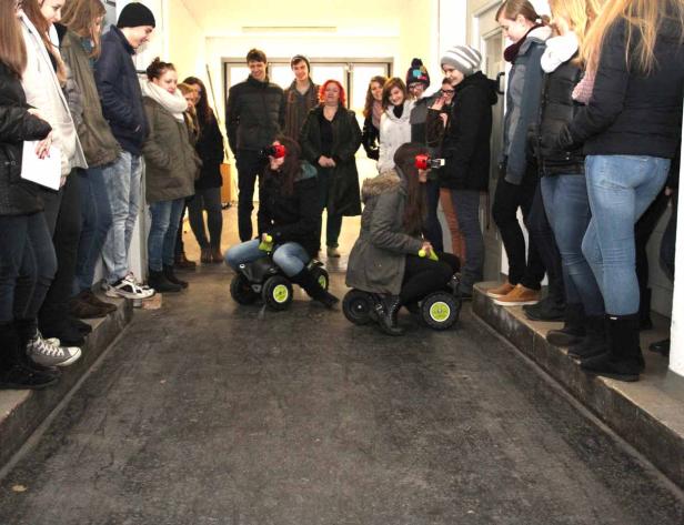 Zwei Mädchen fahren auf kleinen Autos einen Flur entlang, während andere zusehen.