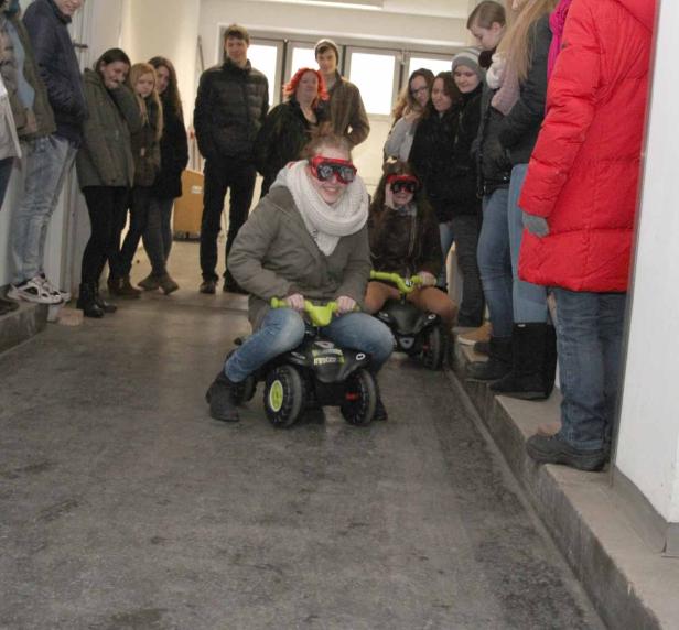 Zwei junge Frauen fahren auf Spielzeugautos einen Flur entlang, während andere zusehen.