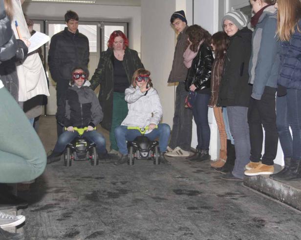 Zwei Mädchen mit Schutzbrillen fahren auf kleinen Autos, während eine Gruppe von Menschen zusieht.