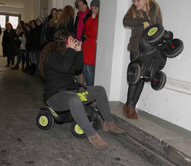 Eine junge Frau fährt auf einem Bobby-Car, während eine andere ein Bobby-Car an eine Wand lehnt.