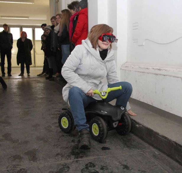 Ein Mädchen mit Schutzbrille fährt auf einem Spielzeugauto einen Flur entlang.