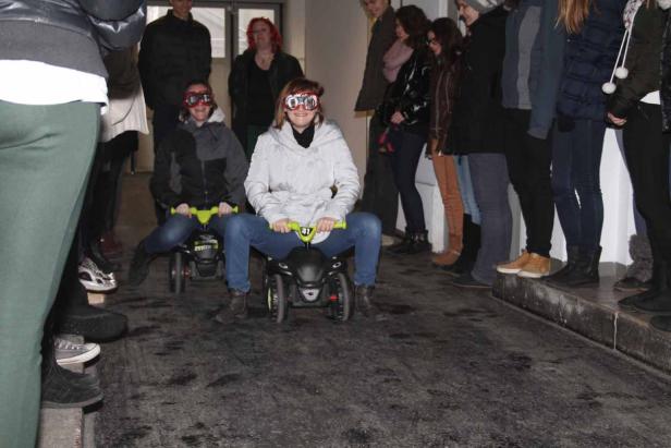 Zwei Frauen mit Schutzbrillen fahren auf kleinen Rutschautos einen Gang entlang.