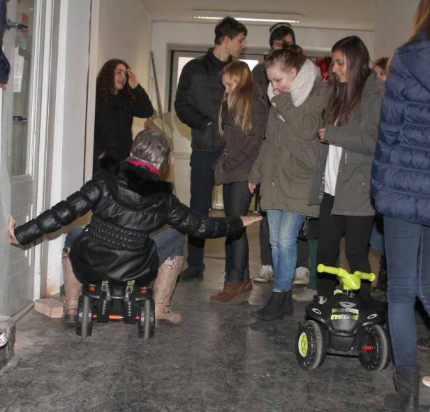 Eine Frau sitzt auf einem Kinderspielzeugauto und balanciert, während eine Gruppe Jugendlicher zusieht.