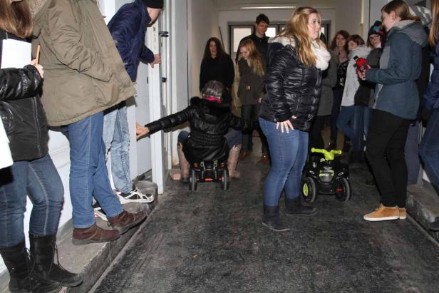 Eine Frau fährt auf einem kleinen Spielzeugauto durch einen Flur, umgeben von Zuschauern.