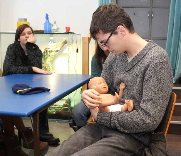 Ein Mann hält eine Übungspuppe für Babys im Arm, während andere Personen zusehen.