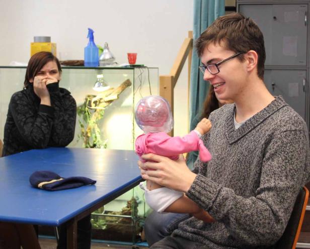 Ein junger Mann hält eine Babypuppe mit einem durchsichtigen Kopf in den Armen, während eine Frau zusieht.