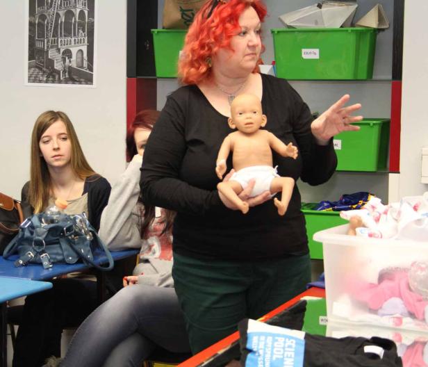 Eine Frau mit roten Haaren hält eine Babypuppe in einem Klassenzimmer.
