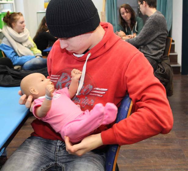 Ein junger Mann hält eine Babypuppe im Arm, während andere Personen im Hintergrund sitzen.