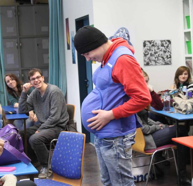 Ein junger Mann trägt einen Schwangerschaftssimulator vor einer Gruppe von Zuschauern.