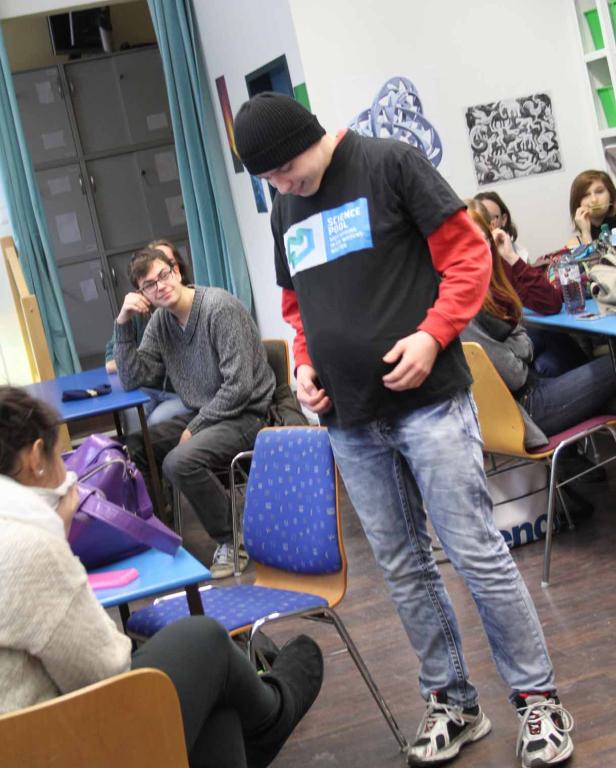Ein Mann mit Mütze und T-Shirt steht in einem Klassenzimmer vor anderen Personen.