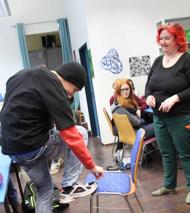 Ein Mann stellt seinen Fuß auf einen Stuhl, während eine Frau und andere Personen im Raum stehen.