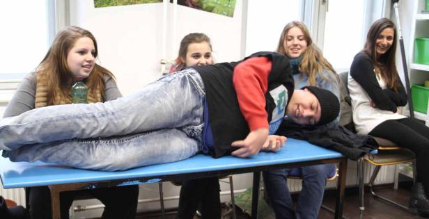 Ein junger Mann liegt auf einem Tisch, während drei junge Frauen um ihn herumstehen und lachen.