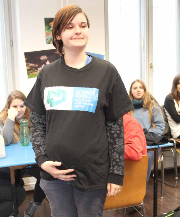 Eine junge Frau mit einem „Science Pool“-T-Shirt steht in einem Klassenzimmer.