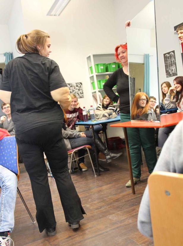 Eine Frau hält einen Spiegel in einem Raum mit mehreren sitzenden Personen.