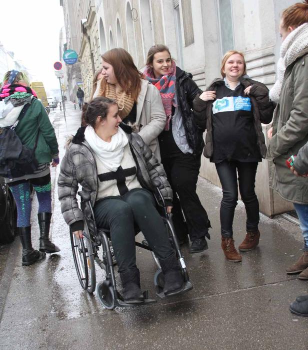 Eine junge Frau im Rollstuhl wird von einer Gruppe junger Leute auf einer nassen Straße begleitet.