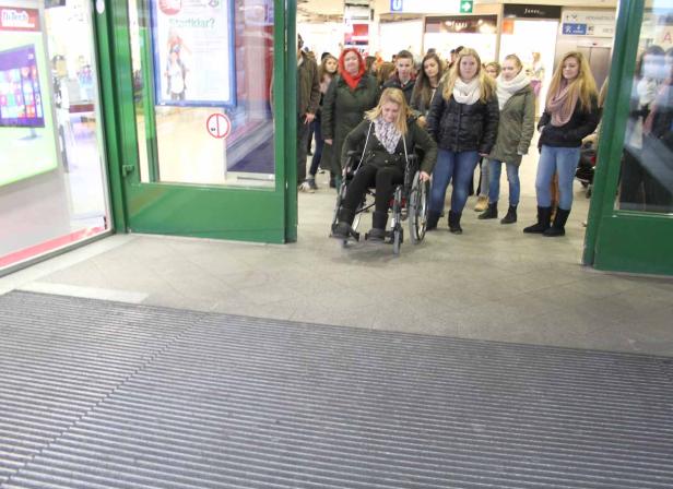Eine junge Frau im Rollstuhl verlässt ein Gebäude, gefolgt von einer Gruppe von Menschen.