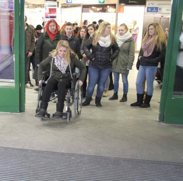 Eine junge Frau im Rollstuhl fährt durch eine Tür, umgeben von anderen Menschen.