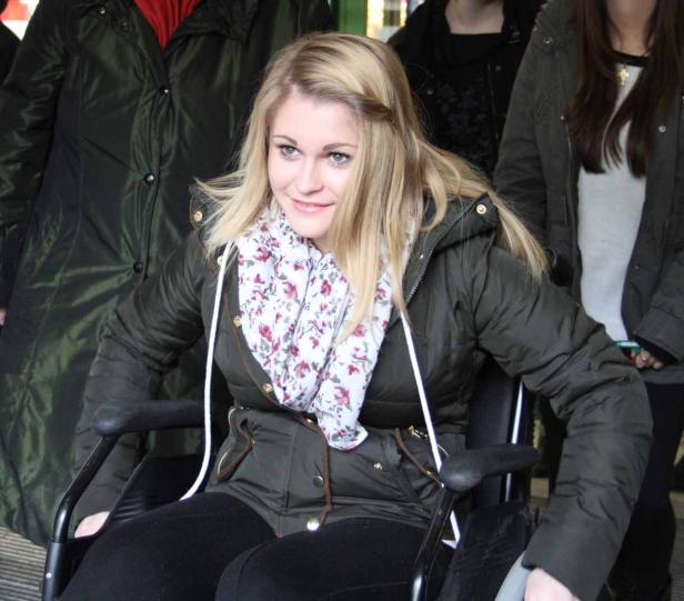 Eine junge Frau mit blonden Haaren sitzt in einem Rollstuhl und lächelt.