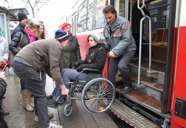 Helfer schieben eine Frau im Rollstuhl in eine Straßenbahn.