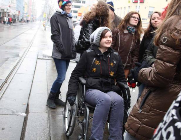 Eine junge Frau im Rollstuhl sitzt inmitten einer Gruppe von Menschen in einer städtischen Umgebung.