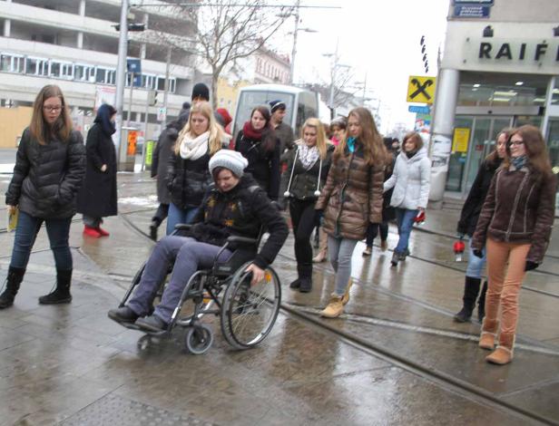 Eine Frau im Rollstuhl fährt auf einer nassen Straße an einer Gruppe von Menschen vorbei.