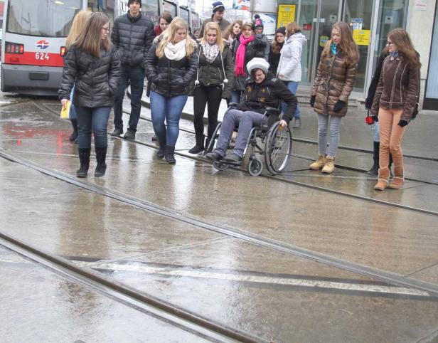 Eine Person im Rollstuhl überquert eine nasse Straße mit Straßenbahnschienen, umgeben von Fußgängern.