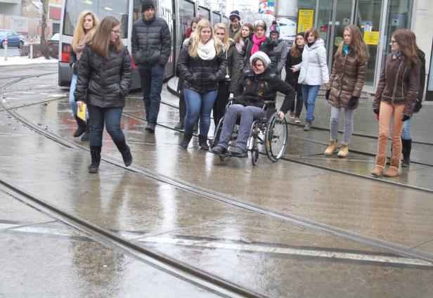 Eine Gruppe von Menschen überquert eine nasse Straße, eine Person im Rollstuhl.