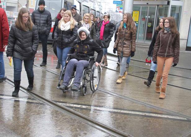 Eine Frau im Rollstuhl fährt über eine nasse Straße mit Straßenbahnschienen.