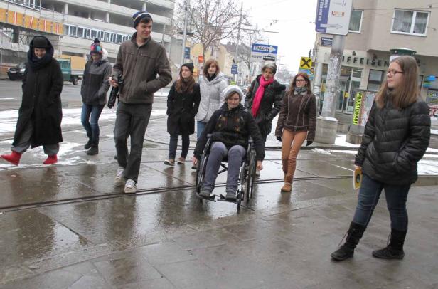 Eine Gruppe von Menschen überquert eine nasse Straße, eine Person im Rollstuhl.