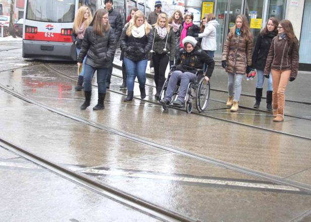 Eine junge Frau im Rollstuhl überquert eine nasse Straße mit Straßenbahnschienen, umgeben von einer Gruppe von Fußgängern.