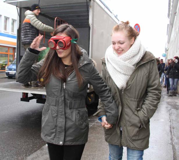 Zwei junge Frauen gehen Händchen haltend die Straße entlang, eine trägt eine übergroße rote Brille.