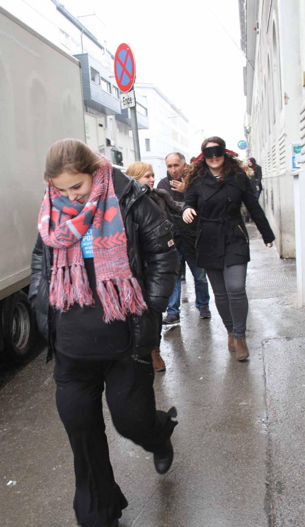 Eine Frau mit verbundenen Augen geht auf einer nassen Straße, gefolgt von anderen Personen.